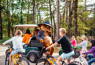 Kinder fahren Fahrrad um ein Maskottchen auf einem Golfwagen im Eurocamping Vessem Ferienpark, Niederlande.