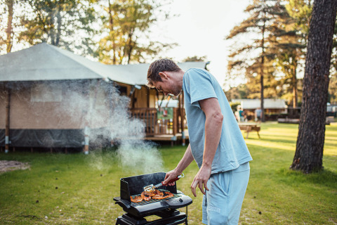 Man grilling food outside at Eurocamping Vessem BV holiday park in North Brabant, Netherlands, with tent behind.