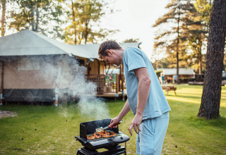 Man grilt buiten eten bij Eurocamping Vessem BV vakantiepark in Noord-Brabant, Nederland, met tent op achtergrond.