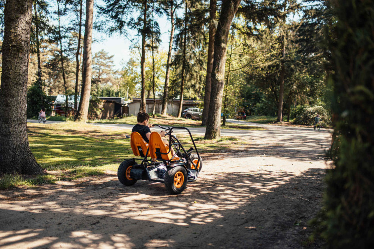 Child rides a four-wheeled pedal go-kart through a sunny forest area at Eurocamping Vessem, Netherlands.