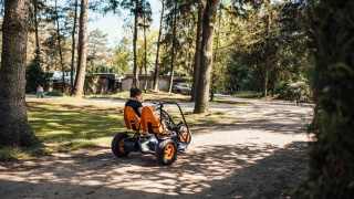 Niño conduce un coche de pedales de cuatro ruedas en un área boscosa y soleada en Eurocamping Vessem.