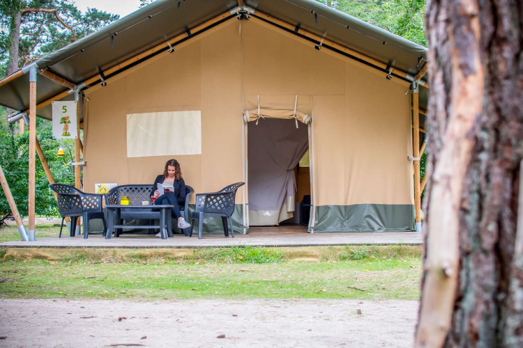Mujer leyendo frente a una tienda Safari en Eurocamping Vessem BV, Países Bajos, rodeada de naturaleza.