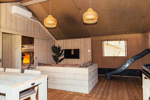 Interior de una tienda safari con paredes de madera, sofá, mesa de comedor y tobogán en Eurocamping Vessem.