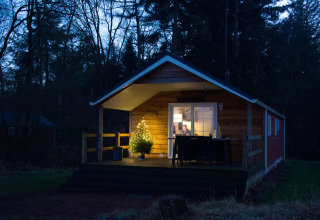 En træhytte med juletræsbelysning på terrassen om aftenen i en skov, set i en feriepark med glamping.
