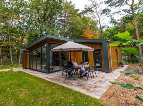 Familie sitzt draußen an einem Tisch vor einer modernen Glamping-Hütte im Ferienpark mitten im Wald.