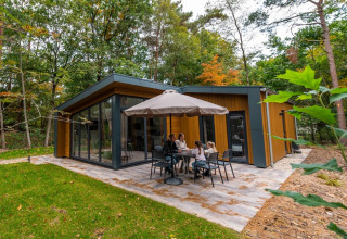 Familie nyder udendørs måltid ved moderne glampinghytte i skoven på feriepark med terrasse og parasol.