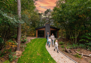 Familie spaziert einen Weg vor einer modernen Hütte im Wald entlang, Sonnenuntergang, Glamping-Urlaub.