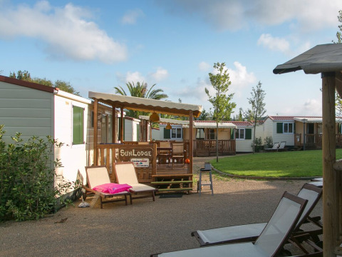 Cozy cabins with wooden decks and loungers in a peaceful, green camping site under a blue sky.