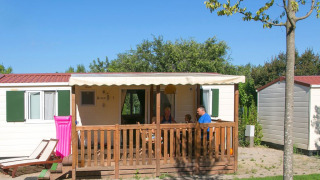 Een familie zit op het terras van een wit chalet met groene luiken en een roze luchtmatras buiten.