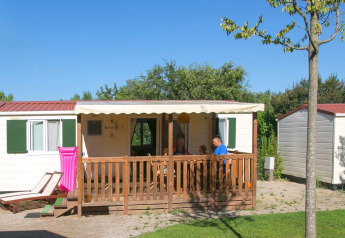Een familie zit op het terras van een wit chalet met groene luiken en een roze luchtmatras buiten.