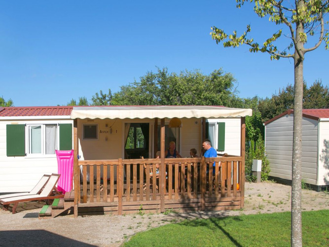 Een gezin zit op de veranda van een witte stacaravan met groene luiken, genietend van het zonnige weer.