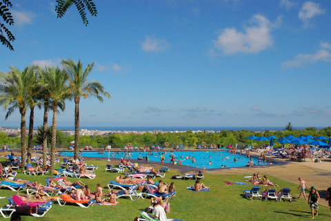 Alojamiento glamping con palmeras, bañistas, gran piscina y vistas panorámicas de la ciudad y el mar.