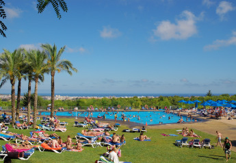 Struttura glamping con palme, bagnanti, grande piscina e vista panoramica su città e mare in lontananza.