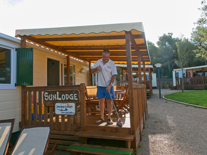 Hombre barriendo el porche de una cabaña SunLodge Redwood en el Camping Vilanova Park en España, día soleado.