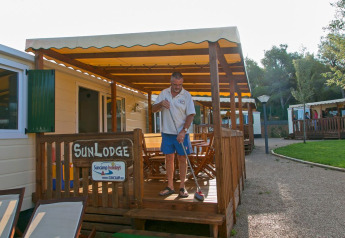 Homme balayant la terrasse d’un bungalow SunLodge Redwood au Camping Vilanova Park en Espagne, journée ensoleillée.