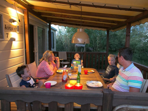 Una familia cena junta en el porche cubierto de una cabaña, rodeada de naturaleza al anochecer.