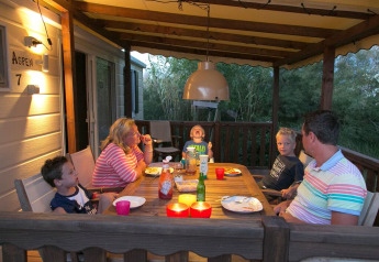 Une famille dîne ensemble sur la terrasse couverte d’un lodge, entourée de verdure en soirée.