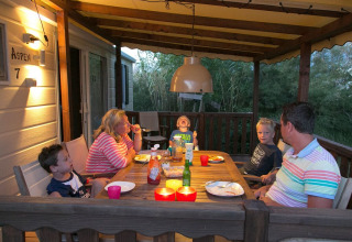 Una familia cena junta en el porche cubierto de una cabaña, rodeada de naturaleza al anochecer.