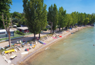 Vista aérea de un camping junto al lago con árboles, caravanas y personas descansando en la orilla rocosa.