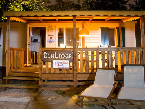 Evening view of SunLodge Maple at Camping Cisano/San Vito in Italy with deck chairs and an illuminated porch.