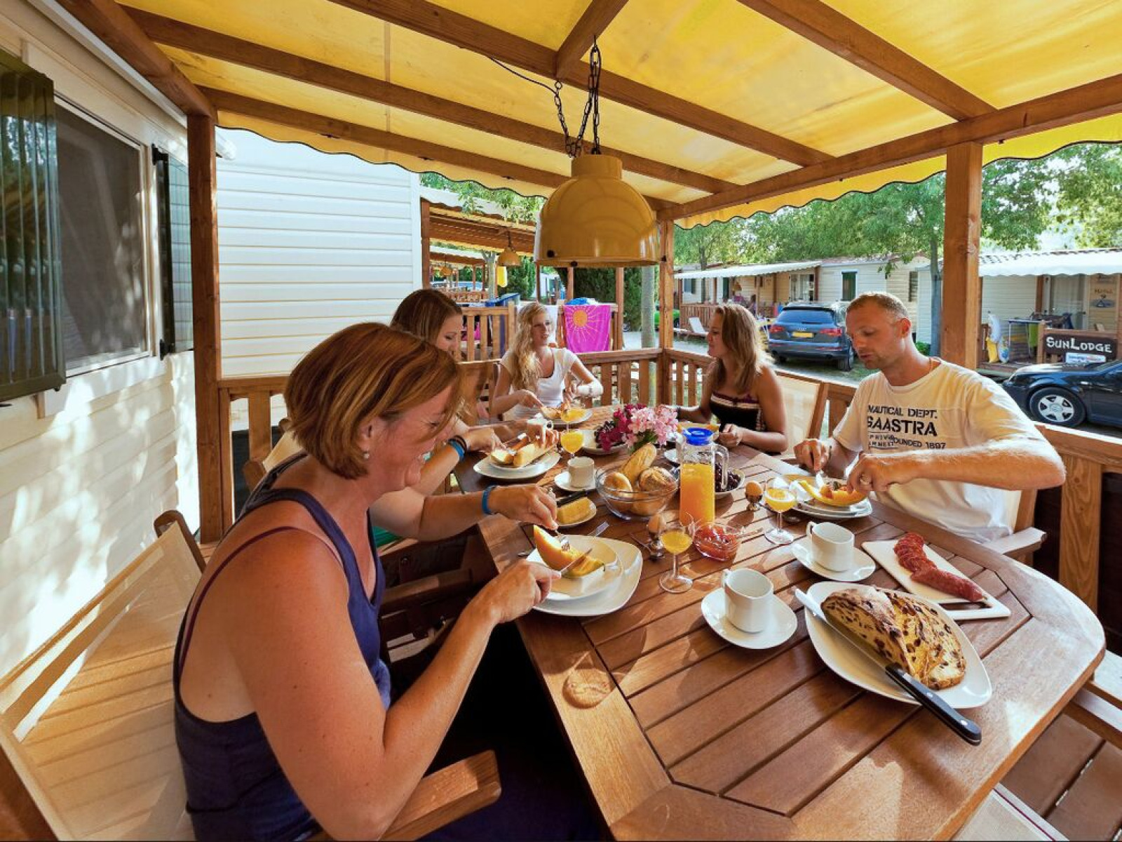 Gezin ontbijten samen op de veranda van een SunLodge Maple, met vakantiehuisjes op de achtergrond.
