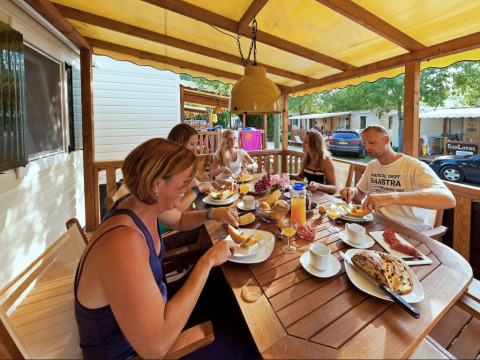 Familia desayunando junta en el porche de una SunLodge Maple, con cabañas de vacaciones alrededor.