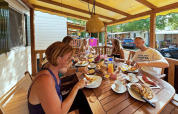 Familia desayunando junta en el porche de una SunLodge Maple, con cabañas de vacaciones alrededor.