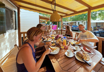 En familie spiser morgenmad sammen på terrassen ved en SunLodge Maple, omgivet af feriehytter.