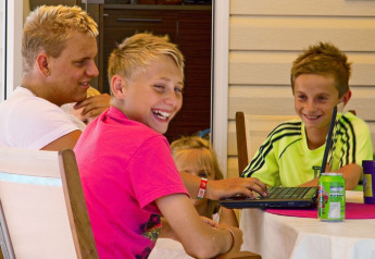 Fire børn slapper af og smiler sammen ved et bord med en bærbar computer i SunLodge Maple, Camping Cisano/San Vito, Italien.