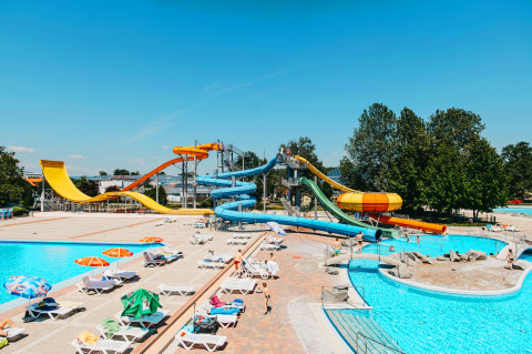 Parco acquatico con scivoli colorati, piscine e lettini sotto gli ombrelloni in una giornata di sole.
