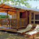 Et SunLodge Jungle teltlodge med en veranda, træmøbler og to liggestole udenfor i skyggen af træer.