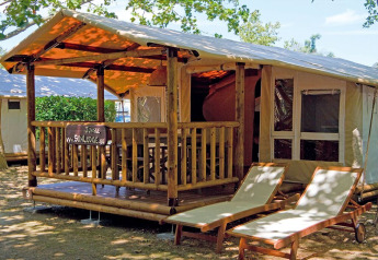 Tenda lodge SunLodge Jungle con veranda, mobili in legno e due lettini prendisole sotto gli alberi.