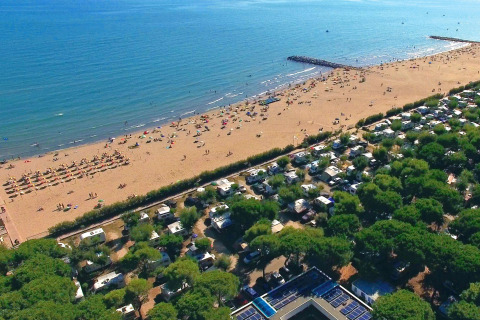 Luchtfoto van een strand met parasols en een camping vlakbij, omringd door dichtbegroeide bomen.