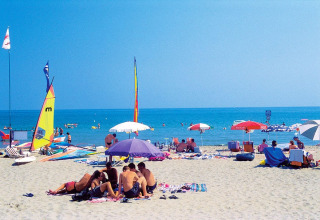 Mensen genieten van een zonnige stranddag met kleurrijke parasols, zeilboten en helderblauwe zee.