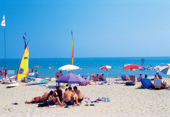 People relaxing on a sunny beach with colorful umbrellas, sailboats, and clear blue sea in the background.