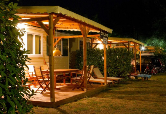 Foto nocturna de un lodge acogedor con terraza de madera, muebles de exterior y luz ambiental cálida.