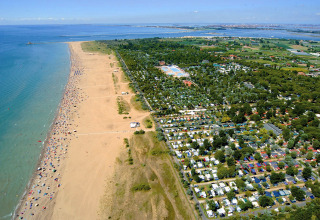 Vista aérea de un camping junto a una amplia playa de arena, con caravanas, árboles y el mar cercano.
