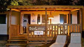 Foto serale della SunLodge Redwood con veranda illuminata, staccionata in legno e due sdraio fuori