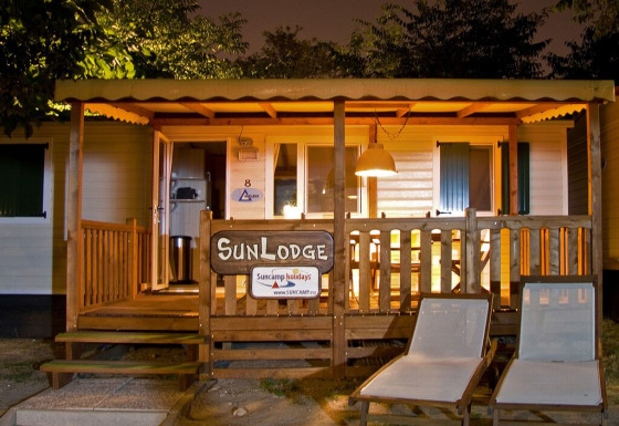 Aftenbillede af SunLodge Redwood hytte med oplyst veranda, træhegn og to loungestole udenfor