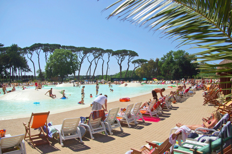 Buitenzwembad bij een lodge met mensen die zonnebaden en zwemmen, omgeven door bomen en ligstoelen.