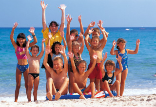 A cheerful group of children and an adult wave on a sandy beach by the sea during a sunny lodge holiday.
