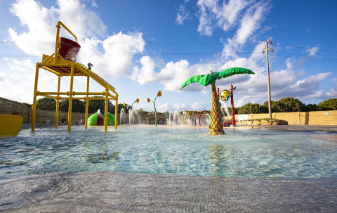 Buitenspeelparadijs met water bij een lodge met kleurrijke speeltuigen, palmbomen en fonteinen onder de zon.