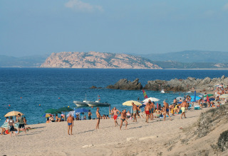 Overvolle zandstrand met parasols, zwemmers en rotsachtige kust op de achtergrond tijdens zomerdag.