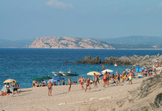 Playa concurrida con sombrillas y bañistas; costa rocosa y mar azul de fondo en un día soleado.