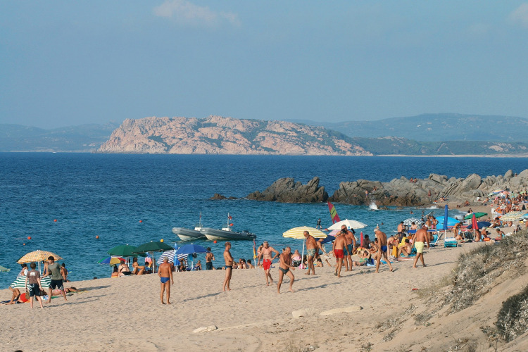 Playa concurrida con sombrillas y bañistas; costa rocosa y mar azul de fondo en un día soleado.