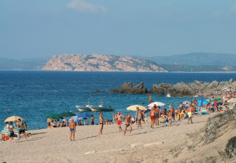 Playa concurrida con sombrillas y bañistas; costa rocosa y mar azul de fondo en un día soleado.
