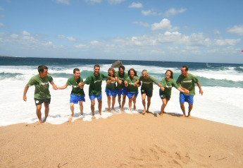 Een groep mensen in groene T-shirts houden elkaars hand vast terwijl ze samen het strand oplopen.