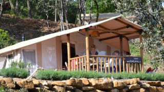 Two people relax on the porch of a SunLodge Safari tent nestled in a wooded hillside with stonework.