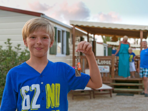 Niño sostiene llaves de SunLodge Redwood en Camping Village Mare Pineta, Italia, familia al fondo.