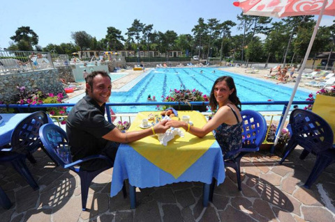 Una pareja sonriente sentada en una mesa cerca de una piscina en un albergue, rodeados de flores y sillas.